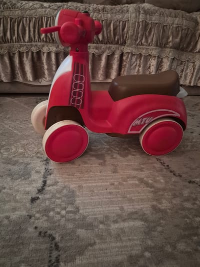 The image shows a red and white toy scooter with a brown seat, resting on a gray carpet. The scooter is the primary subject of the photograph, positioned in the center of the frame. In the background, a plush, tufted bed skirt in a beige or tan color is visible. The floor appears to be carpeted with a patterned design. The lighting suggests an indoor setting. No specific location in the city of Қанабек, Kazakhstan can be determined from the visual information provided.
