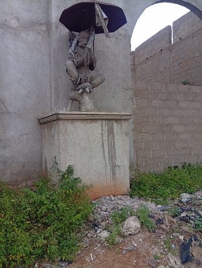 The scene at Itakpe Junction, Nigeria, features a light grey concrete statue of a grotesque, anthropomorphic figure, possibly female, positioned within a concrete architectural recess. The figure, exhibiting prominent breasts, pointed ears, and a wide-open mouth with a distressed expression, crouches on a narrow, elevated concrete base. It holds an open, dark brown umbrella, partially tattered, above its head with its right hand. Tattered fabric or wing-like protrusions are visible on its back. The statue rests on a multi-tiered concrete pedestal.

The architectural setting includes a smooth, light grey concrete wall to the left, which curves into an arch directly above the statue. To the right, an unfinished or damaged wall constructed from grey cinder blocks features another open archway, revealing a bright, overcast sky beyond. The ground in the immediate foreground consists of dirt and gravel, heavily littered with assorted refuse, including plastic bags, paper, and other debris. Patches of vibrant green foliage and weeds grow around the base of the statue's pedestal and across the foreground. Dark, dried liquid streaks are visible on the right side of the statue's concrete pedestal, along with reddish-brown soil at its immediate base. No individuals or active interactions are observed.