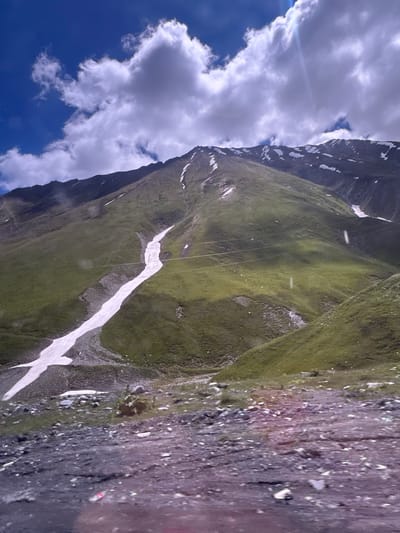 The image depicts a vast mountainous landscape under a bright blue sky with scattered white clouds. A prominent feature is a steep, green mountain slope partially covered with snow, particularly in a long, winding strip that descends from the upper reaches of the mountain. The foreground is rocky and appears to be a road or path with loose stones and gravel. The environment suggests a high-altitude location with sparse vegetation on the mountain slopes. There are no people or specific structures visible that would pinpoint a location within Almasiani, Georgia.