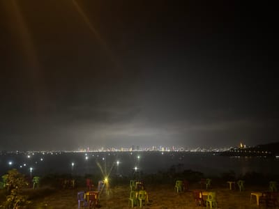 The image captures a nighttime panoramic view from an elevated position within Sơn Trà Ward, Vietnam. The foreground shows an outdoor, unpaved seating area with numerous unoccupied plastic tables and chairs. The chairs are multi-colored, predominantly green, red, purple, and blue, set on an uneven, reddish-brown ground illuminated by warm artificial light sources. One prominent, bright white light source on a short stand is visible in the mid-ground, emitting noticeable lens flares. Beyond the seating area lies a calm body of water, likely a bay, reflecting scattered artificial lights. In the far distance, a dense urban skyline stretches horizontally across the horizon, densely populated with illuminated buildings of various heights, displaying a multitude of colored lights. To the far right of the skyline, on a distant hillside, a large, brightly lit golden structure, consistent with a statue or temple, is visible. The sky above is dark and heavily overcast, with two distinct bright light rays visible in the upper left quadrant. No individuals are present in the scene, and no actions or interactions are depicted.