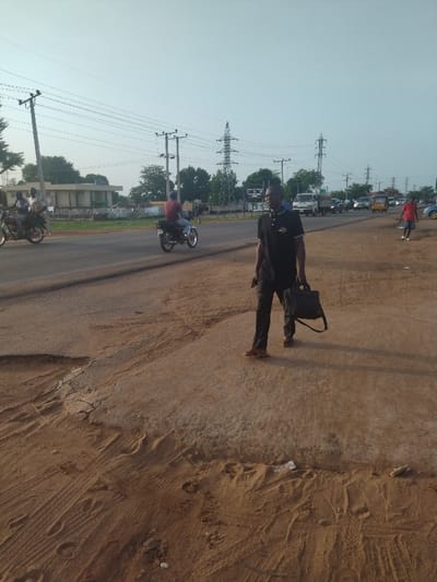 The image shows a man in a black shirt and pants walking on a dirt road in the city of Kanshio, Nigeria. He is carrying a black briefcase. There are power lines in the background and a building on the left side of the image. The man is walking towards the right side of the image. Two motorcycles are parked on the paved road in the background. The ground is covered in dirt and sand. The man is walking on a cracked concrete strip. The road is broken in several places. The road is lined with trees on both sides. The sky is clear and blue.