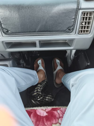 The image depicts the interior of a vehicle, likely a car, from the perspective of someone sitting in the driver's seat. The foreground shows a person's legs and feet. They are wearing light blue trousers and light-colored flip-flops. The left foot is positioned on or near a pedal, possibly the clutch or brake. The right foot is also visible. The floor of the car has a dark textured mat, and a patterned rug with pink and white floral designs is present under the feet.
In the mid-ground, the dashboard is visible. It is made of gray plastic with a black textured panel on top, possibly a dashboard cover. To the right of the dashboard, there is an air conditioning vent. Below the top panel, there are two rectangular storage compartments. A black object, possibly a speaker or a part of the car's console, is partially visible on the right side.
The image does not contain any information that would allow for the specific determination of its location within the city of Buz, Uzbekistan. The visual elements are generic to a vehicle interior.