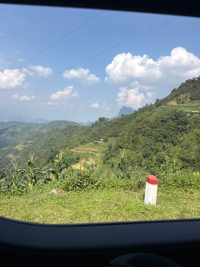 The image shows a view from inside a vehicle, likely a car, overlooking a mountainous landscape in Mậu Duệ Commune, Vietnam. The landscape consists of tiered rice paddies and vegetation-covered hills. In the foreground, there is green grass and corn plants. A white concrete marker with a red top is visible to the right of the frame. The sky is blue with scattered white clouds. Power lines are also visible against the sky.