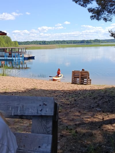 A wide shot captures a serene lake scene in Плюскі сельскі Савет, Belarus on a sunny day. The foreground is dominated by a wooden bench, partially visible, casting shadows on the sandy ground littered with pinecones. To the right, a rustic wooden bench with a backrest sits near the water's edge, occupied by a person facing the lake. In the middle ground, a young person wearing an orange life vest kneels on a paddleboard in the calm, blue water, holding a paddle. The paddleboard is positioned near the sandy shore. To the left, a floating dock with blue pilings extends from a structure that appears to be a small building with a dark roof, and a boat is tied to the dock. The background features a dense line of green trees, beyond which the sky is a clear blue with scattered white clouds. The lake reflects the sky and the surrounding greenery.