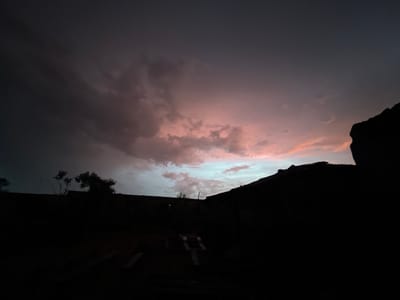 A very dark, low-light photograph captures the sky over Iseyin, Nigeria, at what appears to be either dusk or dawn. The sky is a mix of dark, ominous clouds and patches of pink and blue, indicative of a sunrise or sunset. The foreground is dominated by dark silhouettes of structures, possibly buildings or walls, creating a stark contrast with the sky. There is also the silhouette of some small trees. Details in the foreground are obscured by the low light. The image's composition focuses on the dramatic sky, relegating the cityscape to a mere outline.