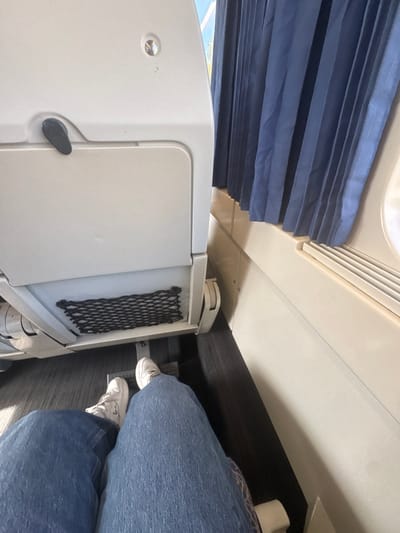 The image depicts an interior view, possibly of a train or airplane, with a blue pleated window shade partially visible at the top left. A white storage compartment or cabinet with a black mesh pocket on its side is situated on the left. The feet and lower legs of a person wearing blue jeans and white sneakers are visible in the bottom right, suggesting someone is seated in the space. The scene provides no information that can be used to deduce if the photo was taken in Trofa, Portugal.