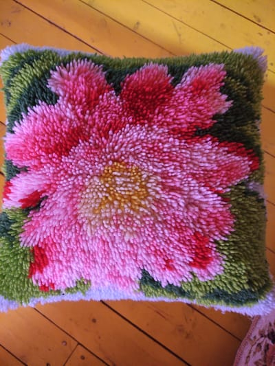 The image shows a close-up, overhead view of a fluffy, textured cushion with a floral design. The cushion is placed on a wooden floor with visible planks and nail heads, appearing to be a light brown or yellow hue. The cushion itself features a large, vibrant pink and red flower at its center, with a yellow and white center. The flower's petals are dense and appear to be made of looped yarn. Surrounding the flower is a green background, also made of the same looped yarn material, creating a textured border. The edges of the cushion are visible, showing a lighter colored fabric underneath the looped yarn. The lighting suggests an indoor setting, possibly a room with natural light. There are no people visible in the image. There is no information in the image that would indicate the location in Kuokunu, Russia.