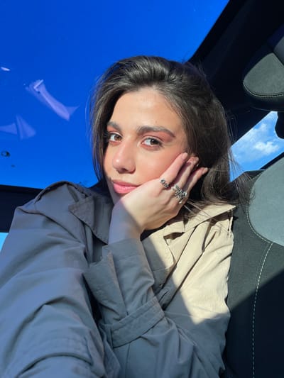 The original image captures a young woman in a close-up portrait, oriented sideways with her head on the left and her body extending towards the right. Describing it as if she were upright, she is seated inside what appears to be a car, looking directly at the viewer with a calm and composed expression. She has long, dark hair, brown eyes, and fair skin. She is wearing a light-colored, possibly beige or olive-toned, trench coat or jacket. Her left hand, adorned with multiple rings featuring intricate, nature-themed designs such as flowers, rests softly against her cheek. Behind her, a dark car seat with light-colored stitching is partially visible. Through the car's window, a bright, clear blue sky with sparse white clouds is visible, indicating a sunny daytime setting. Strong natural light casts distinct shadows across her face and the folds of her clothing. No text is visible in the image. The scene is set in Apaga, Armenia.