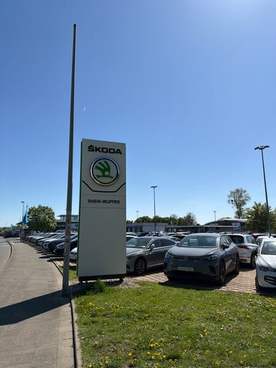 The image depicts the exterior of a Škoda Rhein-Wupper dealership, identified by the tall, vertical sign displaying the Škoda logo and the words "ŠKODA" and "RHEIN-WUPPER." A row of Škoda vehicles are parked along the side of a paved walkway, with a grassy area covered in dandelions adjacent to the cars. The scene is illuminated by bright sunlight under a clear, blue sky. The dealership is located in Leverkusen, Germany.