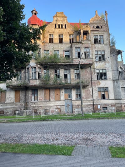 The image shows a large, dilapidated building on a cobblestone street in Sovetsk, Russia. The building appears to be abandoned, with boarded-up windows and peeling plaster. There are several balconies with decorative iron railings, some of which are overgrown with vegetation. The roof is red tile, and there is a red domed turret on the left side of the building. A tall street lamp stands in front of the building. White metal barriers are placed along the front of the building, and grass grows in front of the barriers. The street is paved with cobblestones and a section of grey brick pavement is visible in the foreground. The sky is clear and blue. There are no people visible in the image.
