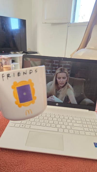 This is a close-up, angled shot of a white laptop open to a scene from the television show "Friends." A woman with blonde hair, appearing to be Phoebe Buffay, is visible on the laptop screen, looking down at something in her hands. In the foreground, partially obscuring the laptop, is a white mug with the "Friends" logo, featuring a purple square framed in yellow. The mug also has the letter "M" in orange. The laptop is placed on a pink, textured surface, likely a blanket or carpet. The background shows a window with white blinds and a dark television screen. The overall impression is a relaxed, indoor setting, possibly a living room or bedroom, where someone is enjoying entertainment. The presence of the "Friends" mug suggests a fan of the show. The lighting appears to be natural, coming from the window, indicating it could be daytime. The overall mood is casual and comfortable.