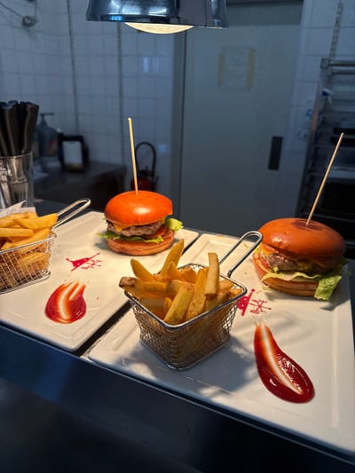This media file captures a close-up view of two burgers and fries served on white rectangular plates, likely in a restaurant setting. The burgers are presented on toasted buns with lettuce and cheese, each secured with a wooden skewer. French fries are served in small, silver wire baskets, positioned beside each burger. Ketchup is artfully smeared on the plates, adding a vibrant red contrast to the food. The background appears to be a kitchen or food preparation area, with tiled walls and metal fixtures, suggesting a professional environment. The lighting is artificial, casting shadows and highlighting the glossy texture of the ketchup. The image is oriented vertically, with the food items arranged in a diagonal line from top left to bottom right. No people are visible, and the overall mood is one of culinary presentation and food service. The location context of Rivesaltes, France, is provided externally and does not offer visual cues within the image itself.