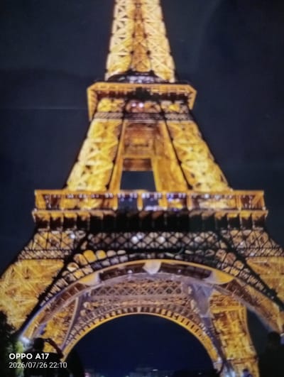 The image shows a close-up, slightly blurry view of the Eiffel Tower at night. The tower is illuminated with golden lights against a dark blue sky. Some architectural details of the lower section of the tower are visible, including intricate latticework and arches. In the foreground, a silhouette of a person can be seen holding up a phone, appearing to take a photograph of the tower. A timestamp "OPPO A17 2026/07/26 22:10" is overlaid on the bottom left corner of the image, indicating it was taken with an OPPO A17 phone on July 26, 2026, at 10:10 PM. The location information that this image is of the Eiffel Tower in the city of Құмөзек, Kazakhstan is incongruous with the subject matter, as the Eiffel Tower is located in Paris, France.