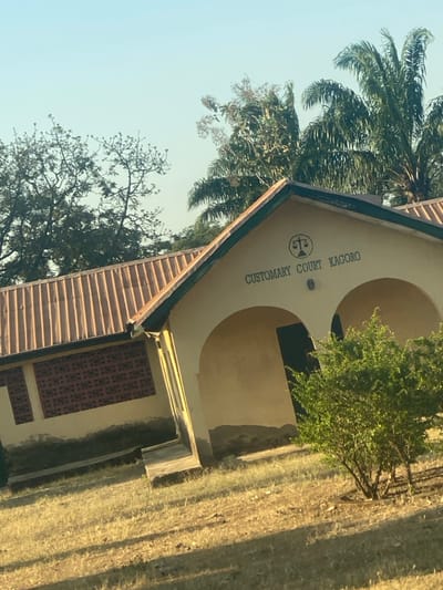 The image captures the exterior of a building identified as the "CUSTOMARY COURT KAGORO," located in Fada, Nigeria. The building is a single-story structure with light, sandy-colored walls and a reddish-brown corrugated metal roof. A symbol of the scales of justice is prominently displayed on the front gable, above the court's name. The facade features two large, arched recessed sections, likely entrances or window alcoves, which appear darker inside. A patterned decorative panel is visible on a lower wall section to the right.
The court building is set outdoors under a clear, light blue sky. To the left and behind the structure, large palm trees and other green trees provide natural vegetation. A smaller, bushy green tree stands to the right foreground of the building. The ground surrounding the building is covered with dry, light-brown grass, suggesting a sunny environment. The scene is brightly illuminated by natural daylight, indicating it is daytime, possibly morning or late afternoon. There are no people present, and no specific activities are taking place in this static view.