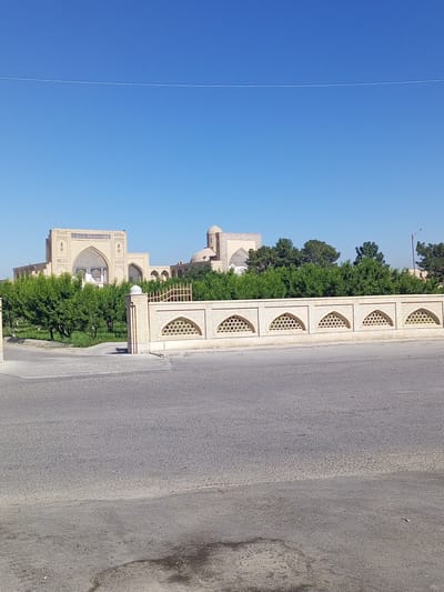 The image captures a wide shot of the historic complex in Shar-Bakr, Uzbekistan, under a clear blue sky. In the foreground, a paved road with some visible staining is present. A decorative brick fence with arched openings runs along the edge of the road, separating it from the complex. Behind the fence, a lush green area with trees is visible. The main subjects of the image are the sand-colored architectural structures of the complex, characterized by arched entrances, domes, and intricate tilework. The buildings appear to be made of brick and stone. The overall environment is dry and sunny, typical of a desert climate. There are no people visible in the scene.