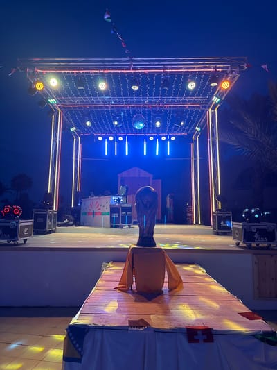 An outdoor stage setup is illuminated with colorful lights and string lights at dusk or night in Taguermess, Tunisia. A DJ booth with a DJ and two people behind it is positioned on the elevated stage. Several speakers and lighting equipment are visible on the stage and in front of it. A large trophy resembling the FIFA World Cup is prominently displayed on a wooden platform in the foreground. Flags of various countries are draped around the platform and hanging above the stage. Palm trees and buildings are faintly visible in the background.