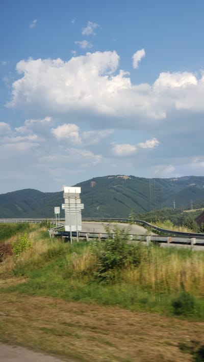 The image depicts a roadside scene with a blue sky filled with white clouds. In the foreground, there is a grassy embankment and a road with a metal guardrail. Several white traffic signs are visible along the roadside. In the background, there are rolling green hills covered with trees. The scene appears to be a landscape view. The location is identified as Turany, Slovakia.