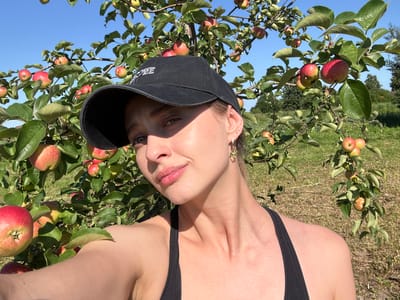A young woman wearing a black baseball cap and a black tank top stands in front of an apple tree laden with ripe red apples. The woman is looking at the camera with a slight smile. The sun is shining brightly, casting shadows on her face. The background is a blur of green leaves and blue sky, with hints of trees and a field in the distance. The scene appears to be outdoors, possibly in an orchard or park. The image has a casual, summery feel.
