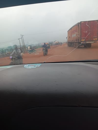 The image depicts a view from inside a vehicle, looking through the windshield at a dusty, overcast outdoor scene. A red and yellow semi-trailer truck is prominently featured on the right side of the frame. The truck is branded with the word "TRANSPORT" in large, dark lettering on its side. The road surface appears to be reddish-brown, suggesting dirt or laterite.

In the background, there is a considerable amount of traffic, primarily consisting of motorcycles and what appear to be tricycles or small three-wheeled vehicles, commonly known as "Keke Napeps" in Nigeria. Several individuals are visible riding these vehicles. There are also utility poles with wires along the side of the road. The atmosphere is hazy or dusty, obscuring distant details.

On the lower part of the windshield, a circular sticker is visible. The sticker contains text and graphics, including "AMAC" (Abuja Municipal Area Council), which suggests the location might be in or around Abuja, the capital city of Nigeria. The overall scene conveys a sense of busy, everyday transportation in a developing urban or semi-urban environment, likely during daytime given the overcast conditions. The presence of dust and the nature of the vehicles suggest a location where such conditions are common, consistent with many parts of Nigeria.