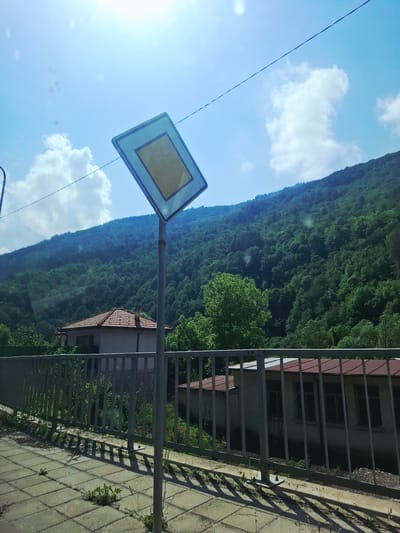 This image captures a vibrant, sunny day in Нареченски бани, Bulgaria, showcasing a mix of natural landscape and residential elements.
In the foreground, a prominent light blue and yellow square road sign, typically indicating a main road or priority, stands on a pole. A thin black power line stretches diagonally across the bright blue sky, which is dotted with scattered white, fluffy clouds. A strong sun flare is visible in the upper left portion of the sky, indicating a sunny time of day, likely afternoon given the shadow direction.
Behind the sign and pole, a steep, densely forested mountain or hill rises, covered entirely in lush green trees, suggesting a natural and unurbanized environment. To the right, a gray metal fence separates a paved sidewalk from a residential building. The sidewalk features square-patterned tiles, and the fence casts long, distinct shadows across it.
Partially visible behind the fence is a house with a reddish-brown tiled roof and light-colored walls. Its windows appear dark, reflecting the light. The scene is devoid of people or any explicit activity, presenting a static snapshot of the location. No discernible text is visible on the sign or elsewhere in the image.