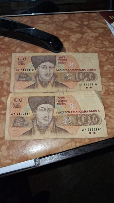 Two Bulgarian 100 Leva banknotes are placed on a brown, patterned surface, possibly a table. Both banknotes feature a portrait of a man wearing a traditional hat and fur-lined collar. The top banknote has the serial number AG 5176710, and the bottom banknote has the serial number AA 7132661. The banknotes are laid out horizontally and are slightly overlapping. The background shows a dark, reflective surface, and part of a black object, possibly a stapler, is visible at the top of the image. There is no indication or information in the image that places the scene in Marikostinovo, Bulgaria.