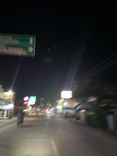 The image depicts a night scene on a street in Crewek, Indonesia. A person is riding a motorcycle on a paved road, illuminated by streetlights and the headlights of passing vehicles, which appear as blurred streaks of light due to motion. Along the sides of the road, there are buildings with illuminated signs, some of which are blurry and difficult to decipher. A green traffic sign with white arrows is visible in the upper left portion of the image. Overhead, power lines are strung across the dark sky. The overall atmosphere suggests a busy street with ongoing activity despite the late hour.