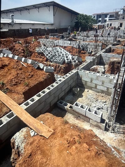 The image depicts a construction site in Ifite Awka, Nigeria, characterized by extensive bricklaying and excavation. The scene is dominated by cinder blocks being used to construct foundations and walls of various structures, indicating ongoing building work. Several construction workers are visible, engaged in their tasks, with some carrying materials. The environment is a mix of disturbed earth from the excavations and the nascent brick structures. The background shows a partially constructed building and a larger, more complete structure, suggesting a developing area. There are also stacks of unlaid cinder blocks, piles of dirt, and some scaffolding. A wooden plank rests on the earth near the foreground.