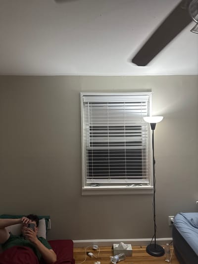 This image captures an indoor scene, likely a bedroom or living area, in Franklin Township, USA, during what appears to be evening or night due to the prominent artificial lighting.
On the left side of the frame, part of a ceiling fan is visible. The walls are painted a light, neutral color, possibly light grey or beige. In the center, a rectangular window is prominent, equipped with white horizontal blinds that are mostly closed, and further secured by white metal security bars. Adjacent to the window, a black floor lamp with a distinctive white, half-moon shaped light fixture stands, providing bright illumination that casts light upwards onto the ceiling and across the room.
To the right of the window, a wooden surface, possibly a nightstand or part of a bed frame, is cluttered with everyday items. These include a white tissue box, several plastic water bottles (one lying on its side), and various charging cables plugged into wall outlets. A dark red blanket covers what appears to be a bed or couch, on which a person is reclining. The person, wearing a dark green shirt, is holding a dark-colored smartphone, with their face mostly obscured by the device, suggesting they are either viewing content or taking a picture. The scene conveys a relaxed, domestic atmosphere. No visible text is present in the image.