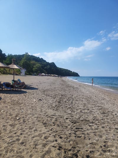The image depicts a wide, pebbly and sandy beach in the Municipal Unit of Melivia, Greece, under a clear blue sky with scattered white clouds. The shoreline is lined with lush green trees and vegetation covering a hillside that stretches along the coast. The sea is a calm, bright blue. In the foreground, two people are relaxing on lounge chairs under a thatched umbrella. Further down the beach, several other umbrellas and lounge chairs are set up, with more people visible in the water and along the shoreline. A lone individual stands at the water's edge in the mid-ground. The overall scene conveys a tranquil beach atmosphere during daytime.