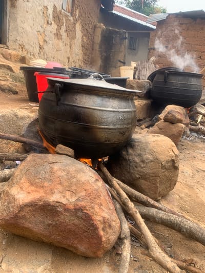 The image depicts an outdoor cooking scene in Zhwarki Mile 10, Nigeria. Two large, black cauldrons are positioned over open fires, with steam rising from one. The cauldrons are made of metal and appear to be old and well-used. They are supported by rocks and firewood. In the background, traditional mud brick buildings with corrugated metal roofs are visible. Several plastic containers, one red and others black, are also present in the scene. The ground is dirt and covered with rocks and branches, indicative of a rural or semi-rural setting. The overall impression is of a functional, rustic cooking arrangement.