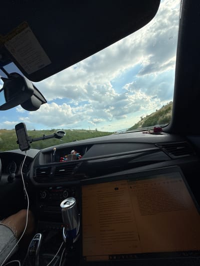The image shows the interior of a car driving through a hilly, grassy landscape under a cloudy sky. The perspective is from the passenger seat looking forward and slightly to the right. A laptop is open on the dashboard, displaying text, possibly in Bulgarian. A red Bull can is secured in a cup holder next to the gear shift. A phone is mounted on the windshield. There are a few small figurines on the dashboard. The sun visor is down. The scene is likely captured on a road in or near Rupkite, Bulgaria, given the landscape.