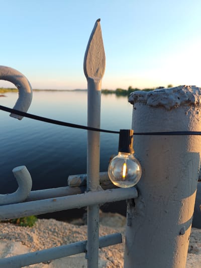 This image, captured in Nikolo-Beryozovka, Russia, displays an outdoor scene illuminated by golden hour lighting, indicating either sunrise or sunset. The primary focus is a weathered grey metal fence structure occupying the foreground and mid-ground. The fence is constructed from painted metal bars, including a prominent, pointed finial on a vertical element slightly left of center, and a thicker, rough-textured vertical post on the far right. A black electrical string light, featuring a single clear, globe-shaped bulb with an illuminated, warm yellow filament, is strung horizontally across the fence, secured to a horizontal bar and wrapping around the thick right post. The background reveals a calm body of water, likely a river or lake, displaying dark blue tones that lighten with reflections. A distant shoreline, densely covered with green vegetation, lines the horizon beneath a clear, light blue sky, which transitions to a warmer hue near the horizon. The ground in the immediate foreground consists of a light-colored, granular material, possibly gravel or concrete, receiving direct sunlight. No people are visible in the scene.