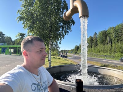 A man is in the foreground of the image, looking to the right. He is wearing a white t-shirt and a chain necklace with a cross pendant. Behind him, a large fountain feature resembles a running faucet, with water spraying into a circular basin. To the left of the man, there is a green bus stop shelter with advertisements for "NESTE." In the background, a road with cars can be seen, bordered by trees and a clear blue sky. The location is identified as the NESTE gas station in Heinola, Finland.