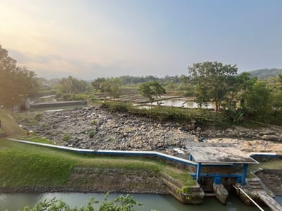 This is a wide-angle, eye-level shot of a rural landscape in Krasak, Indonesia, likely taken in the early morning or late afternoon given the soft, warm lighting. The scene features a small river with a rocky bed, leading towards agricultural fields and lush greenery. In the foreground, a concrete structure with a blue frame and a sloped roof sits over the water, appearing to be a small pumping station or water gate. A paved path with a blue and white railing runs along the grassy bank of the river.
Further into the image, terraced rice paddies are visible, reflecting the sky and surrounded by trees, including palm trees, and other vegetation. Hills covered in trees form the backdrop. The sky is mostly clear with a few wispy clouds and a pale gradient of light. There are no people visible in the frame. The overall impression is of a tranquil, natural environment with evidence of human intervention for water management and agriculture.