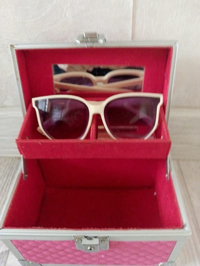 A pair of cream-colored sunglasses with purple lenses sits in a pink, quilted, open jewelry box lined with red velvet. The box has silver trim and clasps. The background appears to be a textured, light-colored wall and a wooden floor. The location is not specified as Prigorognoye, Kyrgyzstan.
