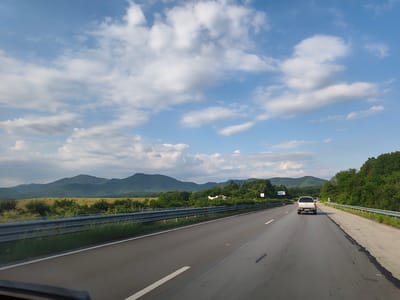 The image depicts a view from a vehicle traveling on a highway, presumably near Osikovitsa, Bulgaria, given the context. The road ahead curves slightly to the right. In the foreground, the asphalt surface of the highway is visible, with white lane markings. A silver pickup truck is traveling in the same direction, ahead of the viewer's vehicle. To the left of the highway, a metal guardrail runs along the edge, separating the road from a grassy embankment. Beyond the embankment, a field with some scattered trees and then rolling hills or mountains covered in vegetation dominate the middle ground and background. The sky above is blue with scattered white cumulus clouds. The overall scene is a landscape view from a moving vehicle on a highway.
