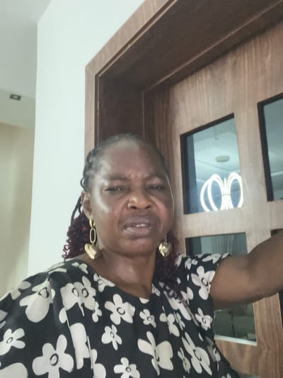 A middle-aged Black woman with braided hair stands indoors, wearing a black and white floral patterned top and gold earrings. She looks towards the camera. Above her is a wood-framed rectangular panel with square windows reflecting a modern light fixture. The background includes a plain white wall. The location in the city of Mopo-Akinlade, Nigeria, cannot be confirmed from the image.