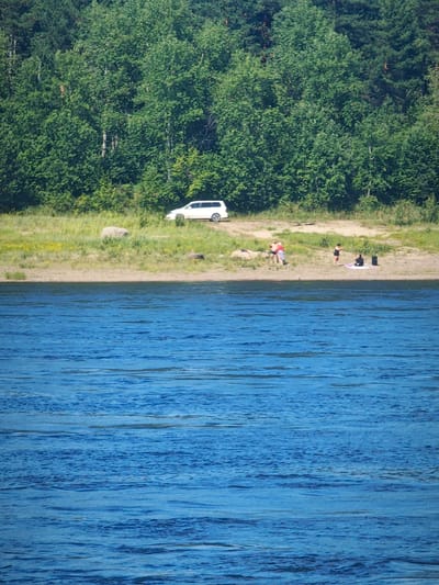 A white van is parked on a grassy area near a sandy riverbank. Several people are visible on the bank. One group appears to be interacting near the van. Further down the bank, a person in dark clothing stands near a mat, and another person sits beside a dark bag. The foreground is dominated by the rippling surface of a blue body of water. The background features a dense forest of green trees.