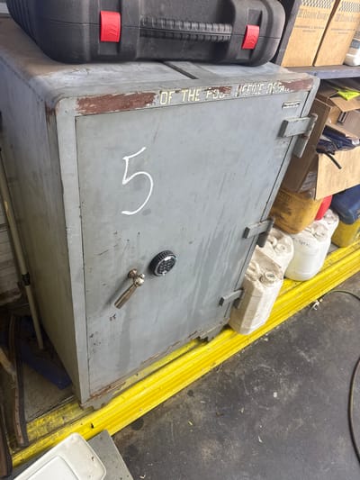 A grey metal safe is positioned on a yellow, ribbed rubber mat. A black toolbox with a red latch rests on top of the safe. The safe has the number "5" painted in white on its front. A dial combination lock and a metal handle are also visible on the safe. To the right of the safe, several plastic containers, some labeled, are stacked. The background includes shelves with cardboard boxes and other items. The floor appears to be concrete. The image is taken from a slightly elevated perspective.