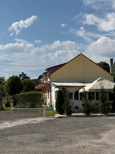 The image shows a building with a yellow facade and a red roof, possibly a restaurant or cafe, situated in a rural setting. In front of the building, there is an outdoor seating area with a large white umbrella providing shade. The area is paved with gravel. A satellite dish is visible on the side of the building. Surrounding the building are trees and green bushes. The sky is blue with scattered white clouds. The scene is lit by natural daylight.