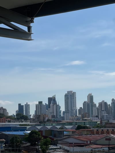 The image depicts an elevated view of the cityscape of Panama City, with the foreground and midground areas consistent with the Curundú corregimiento, looking towards the modern financial district.
The upper left quadrant features a partial view of dark grey metal structural beams, suggesting the vantage point is from an incomplete building or an industrial structure. The sky above is light blue with scattered, thin white clouds, indicating clear daylight conditions.
Below the horizon, the midground displays a densely packed urban landscape. Immediately visible are several light-colored buildings with distinct arched roofs, indicative of industrial or commercial facilities. Further down, a complex array of low-to-mid-rise structures with diverse roof materials – including faded red, blue, and corrugated metallic grey – signifies a residential and mixed-use area. Trees and dense green foliage are interspersed throughout this zone. No individuals are discernible within this section.
In the background, the prominent skyline of modern Panama City rises. Numerous high-rise buildings and skyscrapers, predominantly light in color, are visible. Identifiable structures include a building with "TOWER" marked near its top and the distinctive F&F Tower (El Tornillo), characterized by its spiral design. This arrangement places the observer within or near Curundú, looking eastward or southeastward towards the city's financial center.
