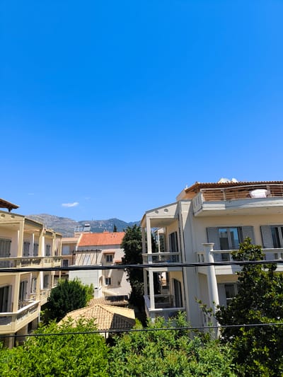 The image depicts a view of residential buildings under a clear blue sky. The buildings are several stories high and appear to be made of light-colored stucco, with balconies and shutters. Some of the buildings have tiled roofs. Green trees and bushes are visible in the foreground and between the buildings. In the background, mountains can be seen, with some structures, possibly antennas, on the peaks. The scene suggests a sunny day in a Mediterranean or similar climate. The presence of a communication tower on one of the mountains might indicate the area is within or near a populated region. The location is identified as the Municipal Unit of Acharnes, Greece.