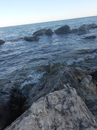 A body of water, likely the sea, is visible with a clear sky above. The water is a deep blue and shows small waves breaking against a rocky shore. The rocks are of various sizes, from small pebbles to large boulders, and are dark gray in color. The image appears to be taken from a low angle, looking out towards the water. There are no people or man-made objects visible in the scene. The overall impression is one of a natural, undeveloped coastline. The location is the Кореизский поселковый совет in Ukraine.