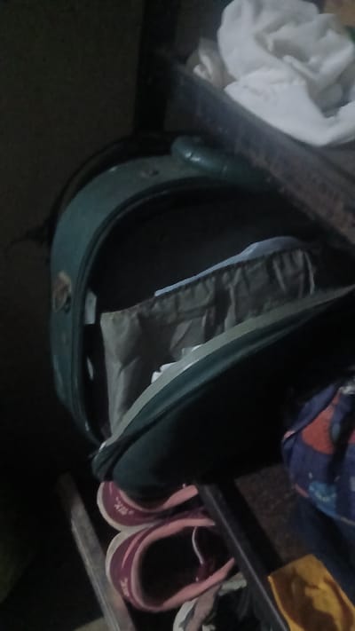 The image shows a partially open green suitcase filled with what appears to be clothing. The suitcase sits beneath a shelf holding other items, including a pair of purple sneakers and various fabrics. The background is dark and indistinct. The image provides no specific evidence to place the scene in Putucual, Venezuela.