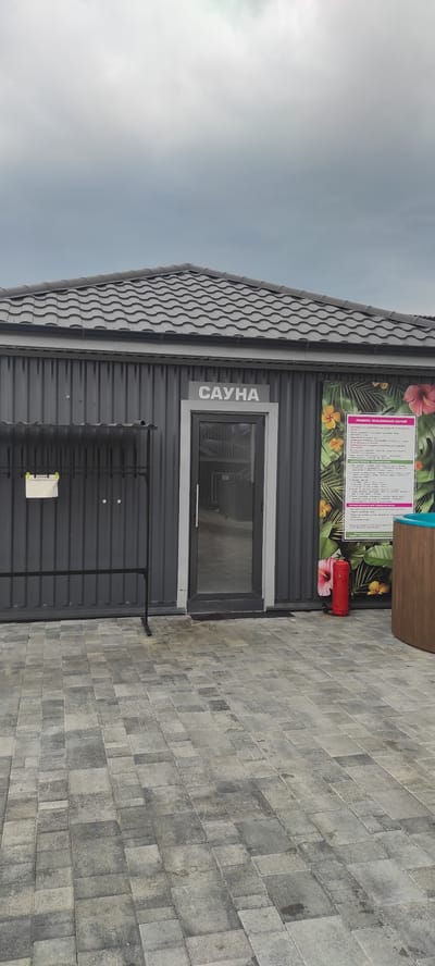 This media file depicts the exterior of a sauna building, likely located in Tulskiy, Russia. The building features a dark, corrugated metal facade and a grey, wavy-patterned roof. Above a rectangular window, a sign reads "САУНА" in large, white, capital letters. To the left of the sign, a colorful mural displays tropical flowers and leaves, adding a touch of vibrancy to the otherwise industrial aesthetic. A bright red fire extinguisher is mounted to the wall near the mural.
The ground in front of the building is paved with grey rectangular tiles, laid in a staggered pattern. The sky above is overcast and pale, suggesting either early morning, late afternoon, or a generally cloudy day. There are no people visible in the scene. The overall impression is one of a functional, perhaps recently constructed, outdoor sauna facility. The presence of the fire extinguisher and the informational posters (partially visible) suggest safety and regulatory compliance. The image is captured from a slightly low angle, emphasizing the building and its surroundings.