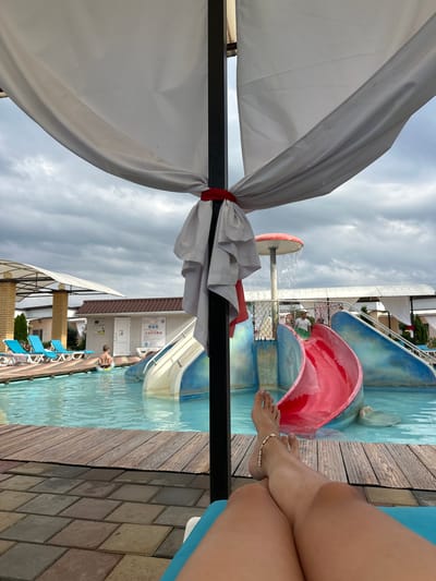 The image depicts a view of a water park from a lounge chair, with the legs of a person visible in the foreground. In the background, there is a swimming pool with blue water, water slides, lounge chairs, and several people. The sky is overcast with clouds. The water park appears to be a leisure facility.