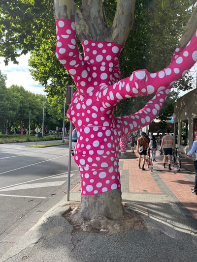 An outdoor, daytime street scene captures a mature tree with its trunk and several lower branches adorned in vibrant hot pink fabric with irregular white polka dots. The fabric is tightly wrapped around the tree, transforming it into a striking art installation.
To the right of the polka-dotted tree, a patterned brick sidewalk stretches into the background. Several people are visible on the sidewalk; some are standing or walking, while others are accompanied by bicycles, suggesting typical urban foot traffic. Beyond the sidewalk lies an asphalt road with white painted lines, possibly indicating a crosswalk or lane markings. More trees, also wrapped in the same polka-dotted fabric, can be seen lining the street in the distance, suggesting this is part of a larger public art display.
The weather appears sunny and clear, casting distinct shadows on the ground and sidewalk. Green foliage from other trees is visible in the background and upper left, contributing to a vibrant, natural setting despite the urban context. The scene evokes a sense of playful artistry within a public space, consistent with areas like Southbank, Australia. No legible text is visible in the image.