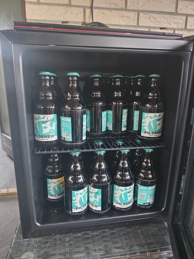 This media file shows an open beverage cooler filled with numerous bottles of Einbecker Pilsener. The cooler is placed outdoors, with a brick wall visible in the background to the left and a woven rattan table and glass surface to the right. The lighting suggests it is daytime. The bottles are arranged in rows, with the distinctive teal and white labels of Einbecker Pilsener clearly visible. The text on the labels reads "Einbecker PILSENER ECHT. EHRLICH." and some also indicate "Alk 4.8% vol." The cooler itself is black with a clear glass door, which is currently open. There are no people visible in the image. The overall scene depicts a collection of chilled beer bottles ready for consumption in an outdoor setting.