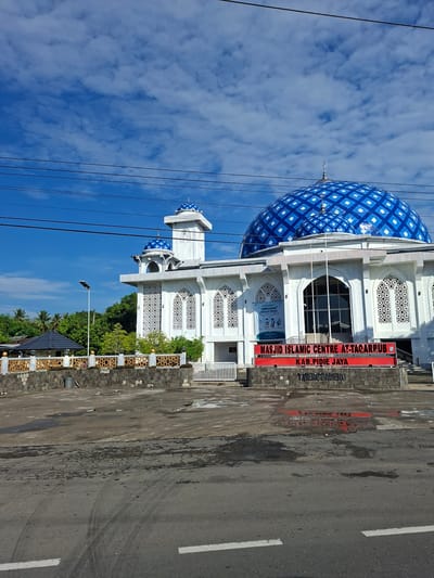 The image shows the Masjid Islamic Centre At-Taqarru in Kab. Pidie Jaya, located in Paya Tringgadeng, Indonesia. The mosque features a prominent large blue and white patterned dome, accompanied by smaller domes and a tall minaret. The building is white with intricate white latticework on its windows. A large red sign at the entrance reads "MASJID ISLAMIC CENTRE AT-TAQARRUB KAB. PIDIE JAYA" and below it "PAYA TRENGGADENG". The mosque is set behind a decorative stone and patterned metal fence. In front of the mosque is a paved area with puddles, indicating recent rain, and a section of road. The sky is blue with scattered white clouds. Trees are visible in the background. There are no people or animals visible in the image. Electrical wires run across the top of the frame.