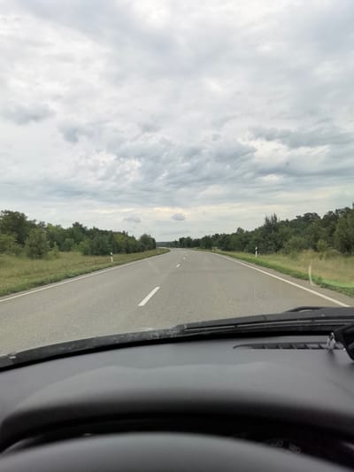 The image shows a view from inside a car traveling on a two-lane road through a rural, forested area. The road is paved with asphalt and has white lane markings. The sky is overcast with gray clouds. The sides of the road are lined with green grass and trees. There are white roadside markers visible along the edge of the road. The dashboard and steering wheel of the car are partially visible in the foreground. The image appears to be taken during the daytime. The location is identified as being in the city of Belogorskiy, Russia.